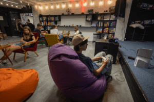 Interiér kultúrno-komunitného dobrovoľníckeho priestoru Hory Doly v Liptovskom Mikuláši, kde mladí ľudia relaxujú a hrajú videohry. Na obrázku sú pohodlné vankúše, herné konzoly a rôzne spoločenské hry na policiach, čo vytvára príjemnú atmosféru pre komunitné stretnutia a aktivity.