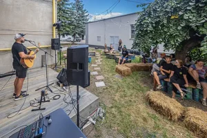 Dobrovoľnícke pozície Živý koncert v kultúrno-komunitnom dobrovoľníckom priestore Hory Doly v Liptovskom Mikuláši, s hudobníkom hrajúcim na gitaru pred publikom sediacim na drevených laviciach a slamenných balíkoch v komunitnej záhrade.